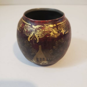 May include: A small, round, red ceramic pot with gold painted designs. The pot has a scene of a house with trees and a gold trim around the top.