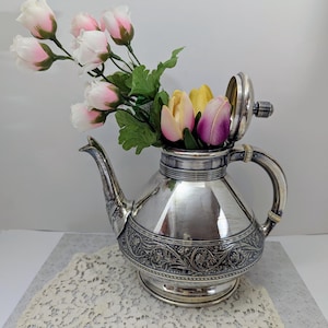 May include: A silver teapot with an ornate design and a hinged lid. The teapot is filled with pink and yellow artificial flowers.