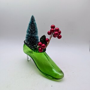 May include: A green glass shoe with a small artificial Christmas tree and red berries inside.