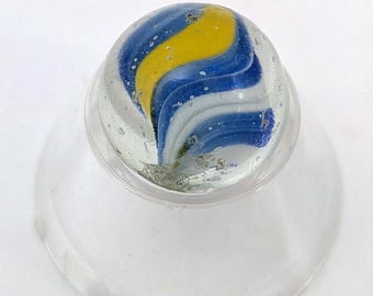 May include: A clear glass marble with a swirl of yellow, blue, and white. The marble is set in a clear plastic holder. The marble's design is reminiscent of a stylized wave.
