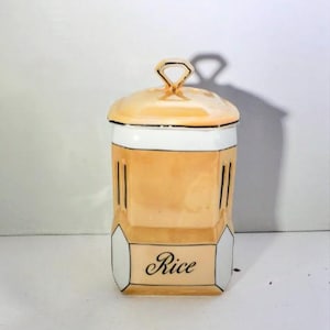 May include: A peach and white ceramic kitchen canister with a matching lid featuring a diamond-shaped handle. The canister has a rectangular body with rounded corners, gold trim accents, and the word Rice printed in black script on the front.