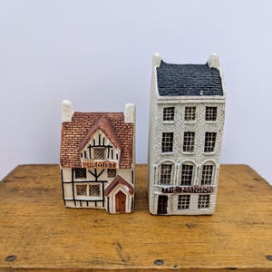 May include: Two miniature ceramic buildings. One is a brown and white Tudor-style building with "CHEMIST" on the sign. The other is a tall, white building with a black roof and "THE MANSION" on the sign. Both are on a wooden surface.