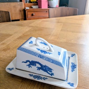 May include: A white ceramic butter dish with a rectangular base and a matching lid featuring a handle. The piece is decorated with blue patterns, including a prominent dragon motif on the sides and floral accents on the lid and base edges.