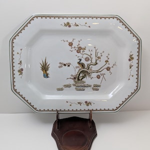 May include: White octagonal serving platter with a detailed peacock and floral design. The platter has a decorative border and is displayed on a wooden stand. The design includes shades of brown, green, and yellow.