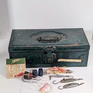 May include: A vintage green metal tackle box with a handle and clasp, labeled "FISHING TACKLE." Assorted fishing lures, hooks, and line are arranged in front of the box. A package of "KADDIS" fishing lures is also visible.