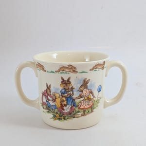 May include: Cream-colored ceramic mug with two handles, featuring a charming illustration of rabbits in a garden setting. The design includes rabbits pushing strollers and holding a balloon. The rim has a repeating pattern of running rabbits.