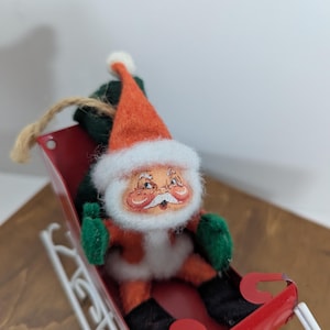 May include: A vintage felt Santa Claus ornament in a red metal sleigh with white trim. Santa is wearing a red hat with white trim, a white beard, and green mittens. He is holding a green bag.