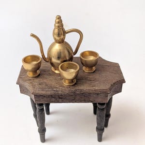 May include: Miniature brass tea set with a teapot and three small cups on a dark wooden table with black legs. The teapot has a bulbous body and a curved spout. The cups are small and round.
