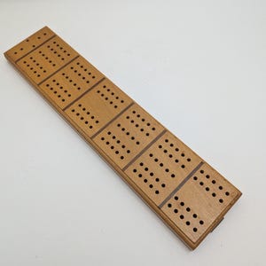 May include: A wooden cribbage board with multiple rows of holes for pegging. The board is rectangular, with a light brown finish and dark brown accents. The holes are arranged in a grid pattern, and the board appears to be unused.