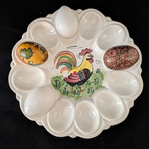 May include: White ceramic egg holder with a colorful rooster illustration. The egg holder has 12 egg-shaped indentations, with three decorated eggs and three white eggs. The rooster is yellow, black, red, and green.