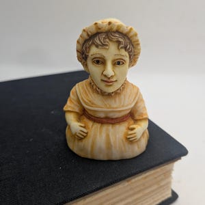 May include: A small, cream-colored figurine of a person wearing a bonnet and a dress with a reddish-brown waistband. The figure has brown hair, detailed facial features, and a ruffled collar, presented in a bust-style design with a rounded base.