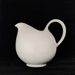 May include: A white ceramic pitcher with a rounded body and a curved handle. The pitcher has a spout for pouring and sits on a small, circular base. The background is black.