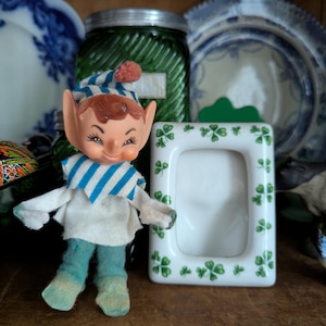 May include: A small, green and white ceramic picture frame with shamrock designs. The frame is empty and has a white background. A small elf doll with a blue and white striped shirt and green pants is standing in front of the frame.