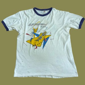May include: A white ringer t-shirt with dark blue trim on the collar and sleeves. The front features a graphic of a yellow electric guitar with blue and red geometric accents and the text "IT'S NOT THAT FUNNY... IS IT?!!" printed above it.