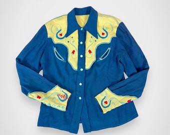 May include: A blue long-sleeved shirt with yellow embroidered details on the collar, cuffs, and yoke. The embroidery features a floral design with red hearts.