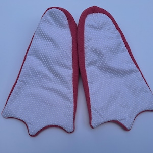 May include: A pair of novelty slippers shaped like duck feet. The slippers have a white, textured interior with a dotted pattern and a red border. The slippers are designed to resemble a duck's webbed feet.