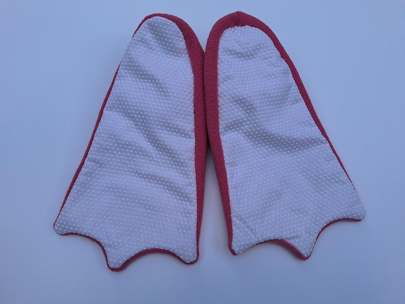 May include: A pair of novelty slippers shaped like duck feet. The slippers have a white, textured interior with a dotted pattern and a red border. The slippers are designed to resemble a duck's webbed feet.