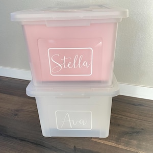 May include: Two clear plastic storage boxes, one pink and one clear, stacked. The pink box has the name "Stella" in white script. The clear box has the name "Ava" in white script. Both boxes have clear lids.