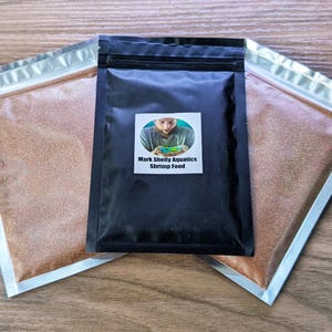 May include: A black resealable pouch with a white label that reads "Mark Shelly Aquatics Shrimp Food". The label also features a photo of a person holding a small bowl of shrimp food. The pouch is filled with a light brown, granular shrimp food.