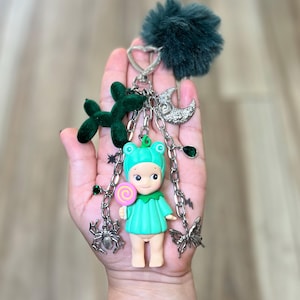 May include: A whimsical keychain featuring a teal frog-themed doll holding a lollipop. The keychain includes a green pom-pom, a velvet balloon dog, and silver charms like a moon, spider, and butterfly. The doll is wearing a teal outfit and a frog hat.