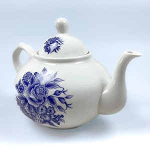 May include: White ceramic teapot with a blue floral design. The teapot has a rounded body, a curved spout, and a handle. The lid is topped with a small knob. The blue floral pattern includes roses and other flowers.