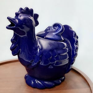 May include: A cobalt blue ceramic rooster-shaped container with a lid. The rooster has an open beak and detailed feathering. The container sits on a wooden tray, with a white background.