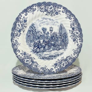 May include: A stack of six white ceramic plates with a blue floral and landscape design. The plates have a scalloped edge and a detailed illustration of a horse-drawn carriage. The plates are stacked on a white surface.