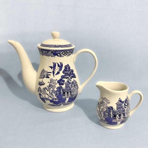 以下が含まれることがあります： 白い陶器のティーポットと、それに合うクリーマー。どちらの作品も、パゴダ、木、鳥が描かれた青い柳模様のデザインが特徴です。ティーポットは長い注ぎ口と小さなつまみ付きの蓋が付いています。クリーマーには取っ手と注ぎ口が付いています。