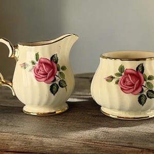 Puede incluir: Juego de jarra de crema y azucarero color crema. Cada pieza presenta un diseño de rosa rosa con hojas verdes y ribete dorado. La jarra de crema tiene asa y pico, mientras que el azucarero es redondo.