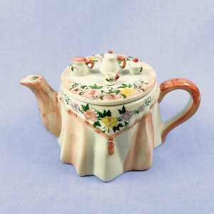Puede incluir: Una tetera de cerámica con un diseño único, con una tapa en forma de mesa puesta para el té. La tapa está decorada con motivos florales y piezas de un juego de té en miniatura. La tetera tiene un asa y un pico de color melocotón.