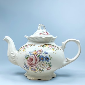 Pode incluir: Uma bule de cerâmica branca com tampa e base recortadas, decorado com um padrão floral colorido de rosas cor-de-rosa, margaridas azuis e flores amarelas. O bule inclui uma pega curva, um bico moldado e detalhes de acabamento dourado.