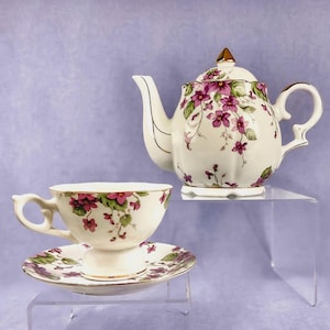 Puede incluir: Un juego de té vintage con tetera, taza y platillo. El juego de porcelana blanca está adornado con un estampado floral de flores moradas y hojas verdes. Los bordes dorados acentúan los bordes del juego, añadiendo un toque de elegancia.