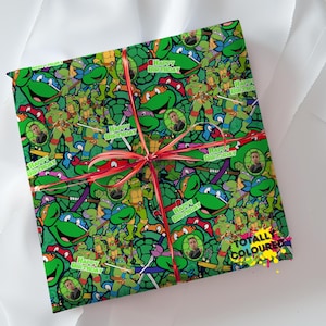 May include: A square gift box wrapped in green Teenage Mutant Ninja Turtles patterned paper featuring the text Happy Birthday and small circular photo inserts, tied with a thin red ribbon. The bottom right corner displays the text Totally Coloured.