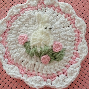 Puede incluir: Un salvamanteles de ganchillo hecho a mano, blanco y rosa, con un diseño de conejito y flores. El salvamanteles presenta un conejito blanco con ojos negros, rosas rosas y hojas verdes. El borde festoneado es blanco y rosa.