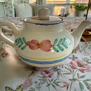 Puede incluir: Una tetera de cerámica blanca con un diseño pintado a mano de una flor rosa central con centro amarillo, flanqueada por hojas verdes. La base está decorada con una banda horizontal amarilla y una línea ondulada azul, con una tapa a juego.