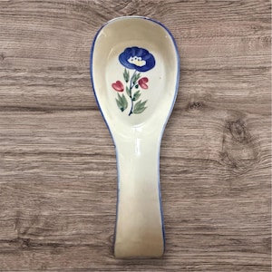 Puede incluir: Un reposacucharas de cerámica de color crema con un borde azul. El centro presenta un diseño floral pintado a mano que consiste en una flor azul grande, dos pequeños capullos rojos y hojas verdes sobre un tallo. Forma ovalada alargada.