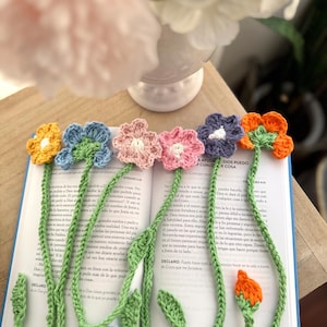 Puede incluir: Marcadores de libros de flores de ganchillo en amarillo, azul, rosa, morado y naranja, con tallos y hojas verdes, colocados en un libro abierto. Los marcadores son hechos a mano.