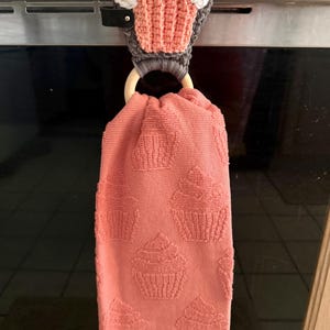 Puede incluir: Un paño de cocina de color coral con un patrón de cupcake cuelga de un soporte de ganchillo con forma de cupcake. El soporte presenta una base gris, un cupcake coral y glaseado blanco con chispas rosas y amarillas. Un anillo de madera asegura la toalla.