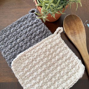 Puede incluir: Dos agarraderas de ganchillo hechas a mano en gris y crema, con un patrón de tejido texturizado. Se adjunta un pequeño lazo para colgar. También se ven una cuchara de madera y una planta en maceta con follaje verde.