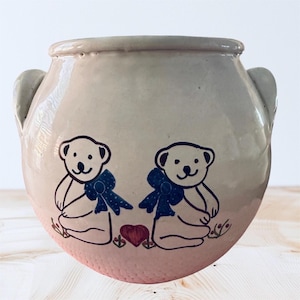 Puede incluir: Olla de cerámica beige con dos asas y cuerpo redondeado. La olla presenta un diseño dibujado a mano de dos osos de peluche con lazos azules y un corazón rojo. Los osos están sentados y la olla tiene un acabado brillante.