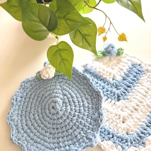 Puede incluir: Dos posavasos de ganchillo hechos a mano. Uno es azul claro liso con un borde festoneado y una pequeña flor blanca. El otro es blanco y azul claro con un borde festoneado. Hojas verdes en el fondo.