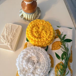 Puede incluir: Un cepillo para fregar con mango de madera y cerdas blancas se encuentra junto a una pastilla de jabón blanca con un diseño de lavanda. Dos estropajos de ganchillo, uno amarillo y otro blanco, descansan sobre un paño de cocina con estampado de limones y una ramita de romero.