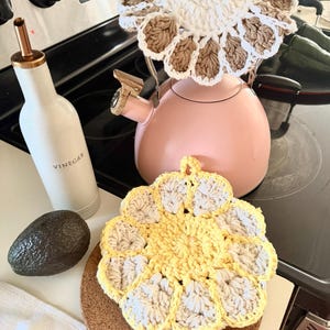 Puede incluir: Una tetera rosa con una cubierta de ganchillo y un salvamanteles de ganchillo amarillo y beige. También se ve una botella blanca con la etiqueta "Vinagre" y un aguacate. Los artículos están sobre una encimera negra.