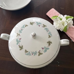 Puede incluir: Fuente de cerámica blanca con tapa y asas, decorada con una corona de hojas verdes y azules y bayas moradas. Un cuenco a juego es visible en el fondo. Una servilleta rosa con flores blancas está a la derecha.