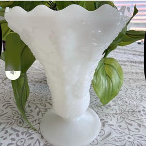 Puede incluir: Un jarrón blanco de estilo vintage con un borde ondulado y una base de pedestal. El jarrón tiene una superficie texturizada con un patrón floral. El jarrón está hecho de vidrio blanco opaco y probablemente se utiliza con fines decorativos.