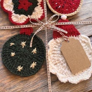Puede incluir: Un conjunto de cuatro adornos de ganchillo atados con una cinta beige. La colección incluye una estrella roja y blanca, una pieza circular roja, un adorno circular verde oscuro con estrellas bordadas y una pieza blanca con una parte superior roja y una etiqueta marrón en blanco.