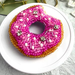 Puede incluir: Un broche con forma de d&oacute;nut elaborado con intrincado trabajo de cuentas. La parte superior presenta cuentas de semillas de color rosa vibrante, acentos de cristal transparente y cuentas cil&iacute;ndricas rosas. La base est&aacute; cubierta de cuentas de color &aacute;mbar transl&uacute;cido, creando una apariencia texturizada y tridimensional con un detalle de mordisco.