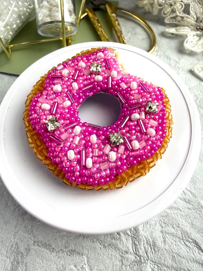 Puede incluir: Un broche con forma de d&oacute;nut elaborado con intrincado trabajo de cuentas. La parte superior presenta cuentas de semillas de color rosa vibrante, acentos de cristal transparente y cuentas cil&iacute;ndricas rosas. La base est&aacute; cubierta de cuentas de color &aacute;mbar transl&uacute;cido, creando una apariencia texturizada y tridimensional con un detalle de mordisco.