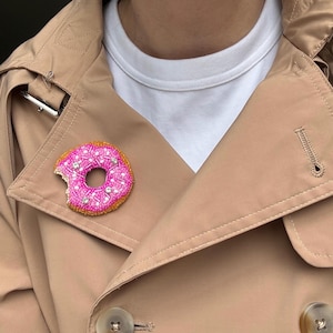 May include: A beaded donut brooch featuring a bright pink frosted top with white and clear crystal sprinkles, a golden brown cake base, and a small bite mark on the left side, pinned to the lapel of a tan trench coat over a white shirt.