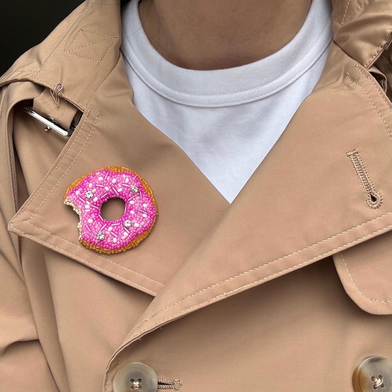 May include: A beaded donut brooch featuring a bright pink frosted top with white and clear crystal sprinkles, a golden brown cake base, and a small bite mark on the left side, pinned to the lapel of a tan trench coat over a white shirt